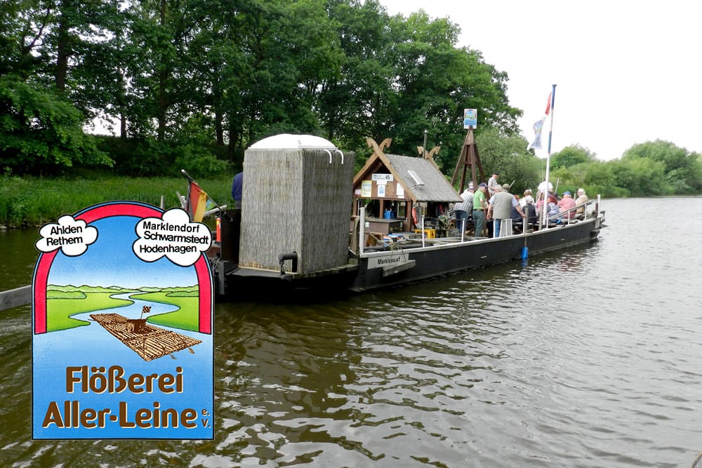 Ein Holzfloß mit mehreren Personen und einer hüttenähnlichen Konstruktion schwimmt auf einem Fluss in der Nähe grüner Bäume. Im Vordergrund steht ein illustriertes Schild mit der Aufschrift 