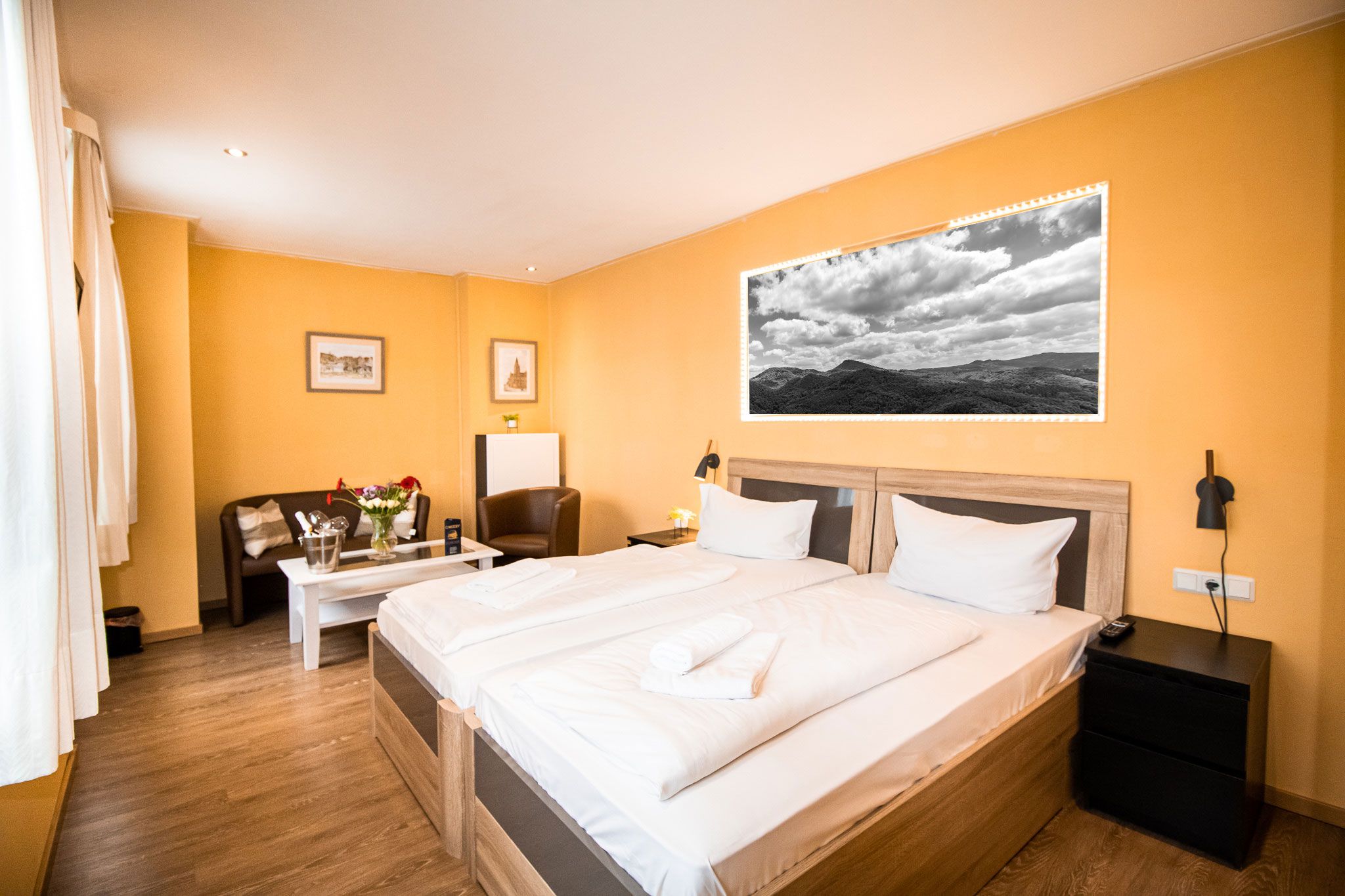 Ein gemütliches Hotelzimmer mit gelben Wänden, einem Doppelbett mit weißer Bettwäsche, Nachttischen, einem kleinen Sofa und einem Couchtisch mit Blumen und einem großen Schwarz-Weiß-Foto über dem Bett.