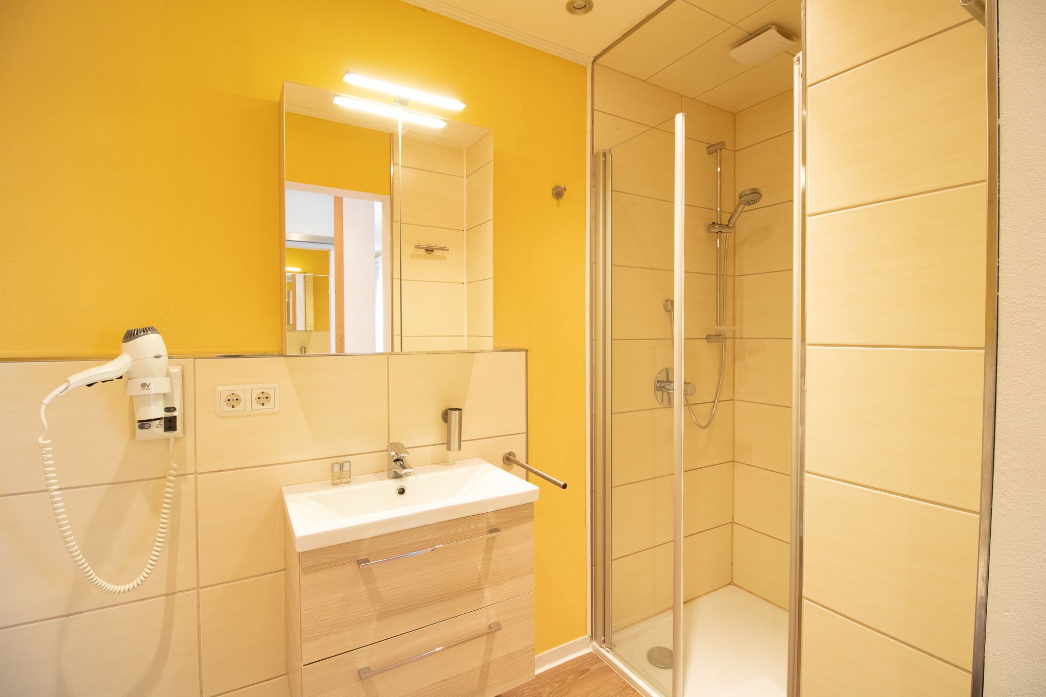 Modernes Badezimmer mit gelben Wänden, einem Spiegel über einem Waschbecken mit Unterschrank, einem eingebauten Haartrockner und einer verglasten Dusche mit Regenbrause und beigen Fliesen.