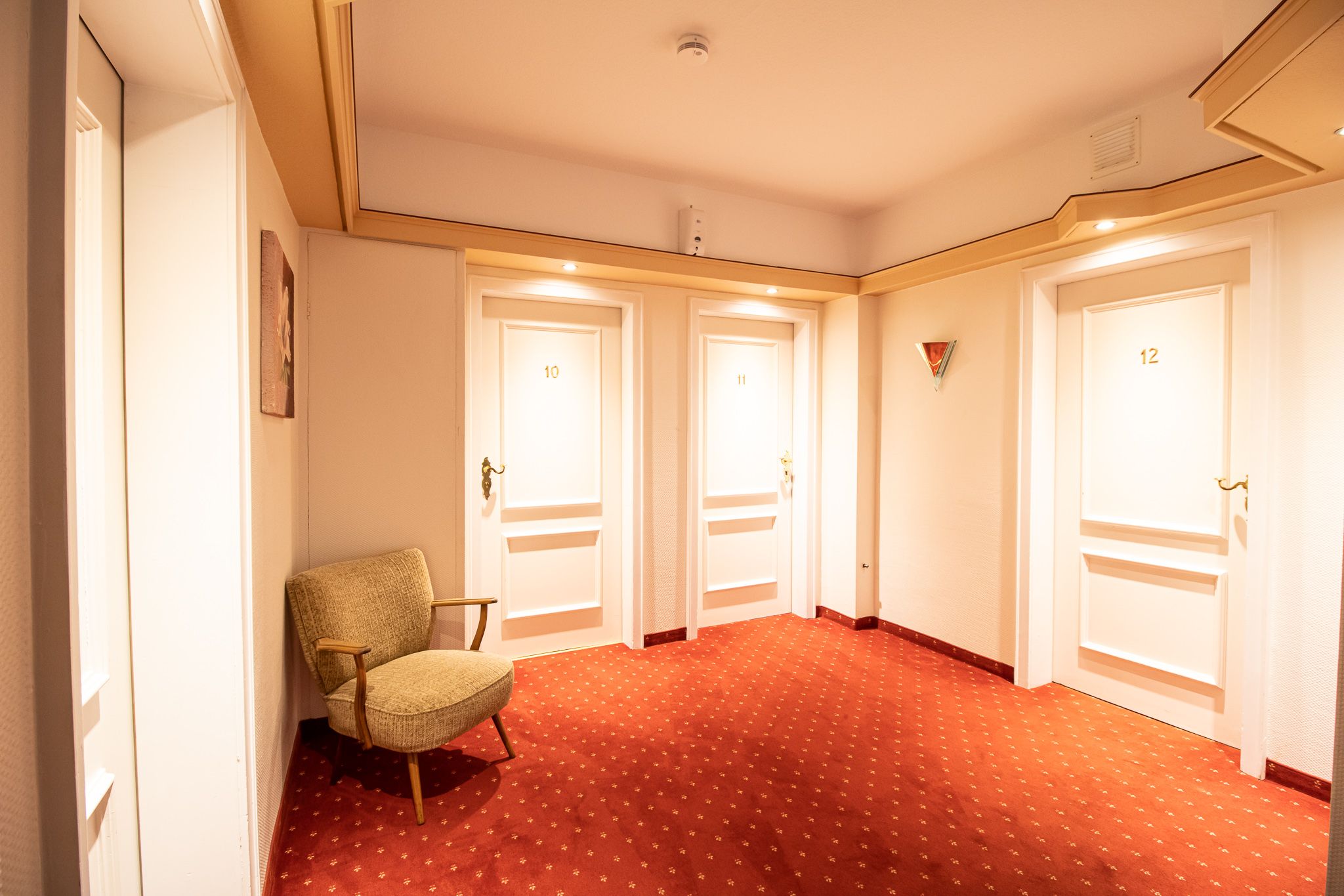 Ein Hotelflur mit rotem Teppich, cremefarbenen Wänden und drei weißen Türen mit den Bezeichnungen 10, 11 und 12. Auf der linken Seite steht ein gepolsterter Sessel, und Deckenleuchten erhellen den Raum.