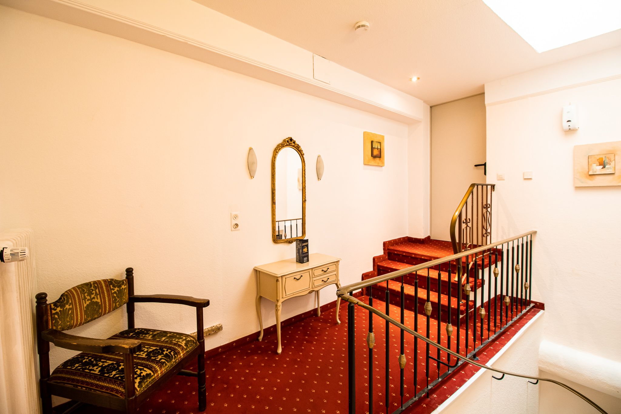 Ein kleiner Flur mit rotem Teppich, einem schwarz-gold gepolsterten Stuhl, einem cremefarbenen Schminktisch mit einem goldgerahmten Spiegel und zwei gerahmten Bildern an weißen Wänden neben einer Treppe mit schwarzem Geländer.