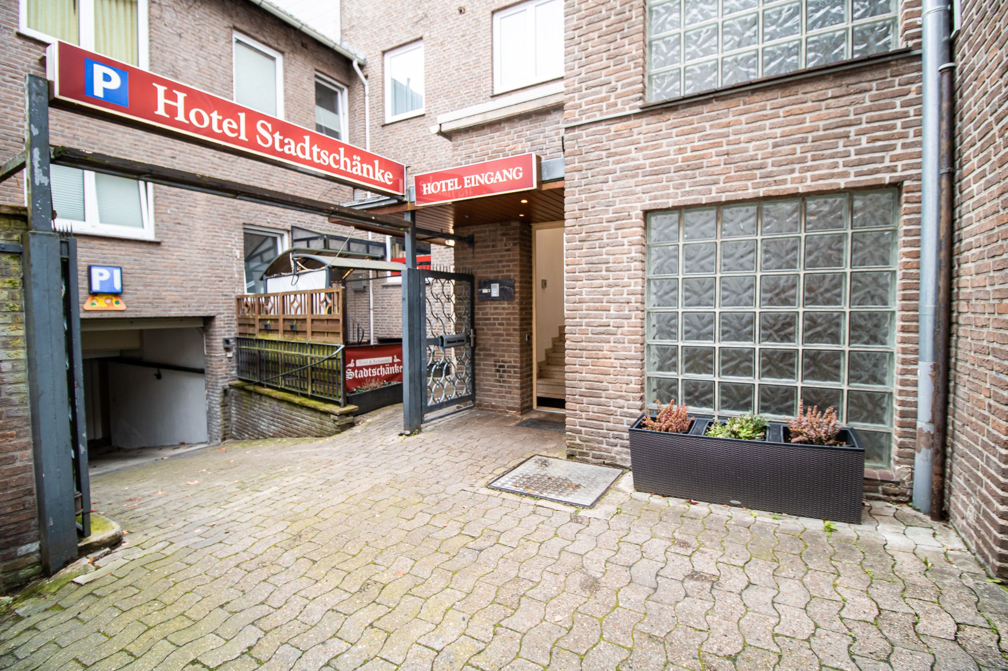 Eingang zum Hotel Stadtschänke, mit einem Schild darüber, Backsteinmauern, Glasbausteinfenstern, einer Rampe, die zur Tiefgarage führt, und einem Pflanzkübel mit kleinen Pflanzen auf einem gepflasterten Innenhof.