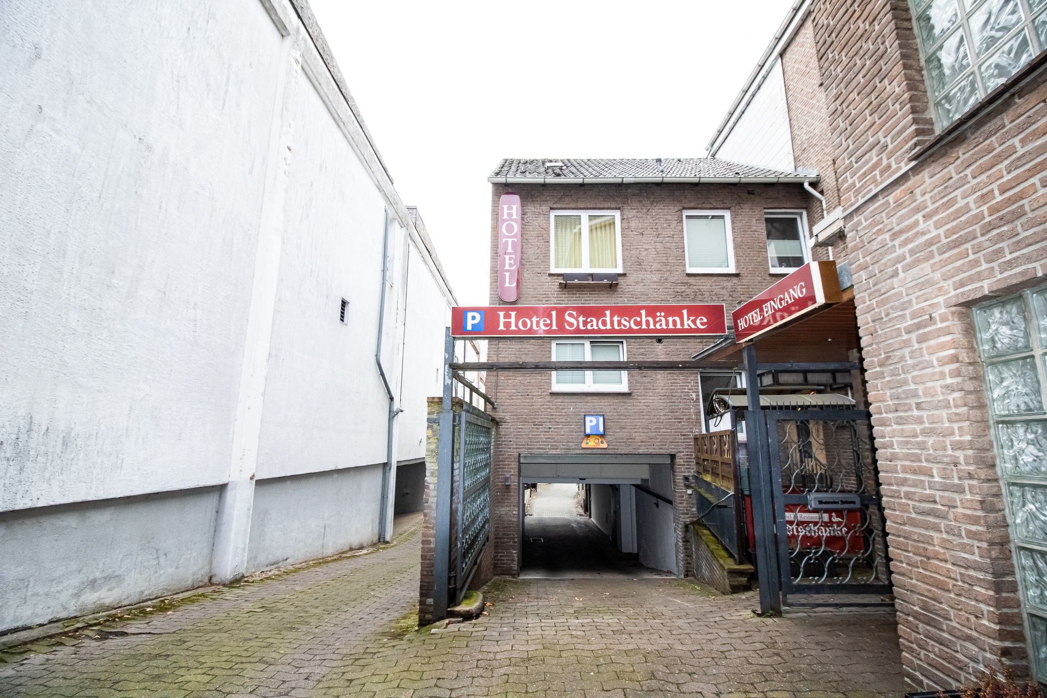 Eine schmale Gasse führt zu einer Tiefgarageneinfahrt für das Hotel Stadtschänke, mit Schildern für Parkplatz und Hoteleingang an einem Backsteingebäude. Die Umgebung besteht aus weißen und braunen Mauern und einem gepflasterten Gehweg.