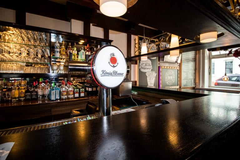 Eine gut beleuchtete Bar mit einem König-Pilsener-Zapfhahn in der Mitte, Regalen mit Spirituosenflaschen und Gläsern hinter der Bar und einem großen Fenster auf der rechten Seite, das die Straße draußen zeigt.