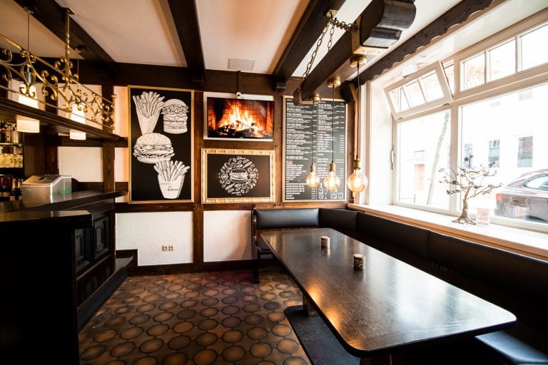 Ein gemütliches Restaurant mit einer schwarzen gepolsterten Bank, einem dunklen Holztisch, Wandbildern von Burgern und Pommes frites, einem Kaminbild, Hängelampen und einer großen Speisekarte an einem hellen Fenster.