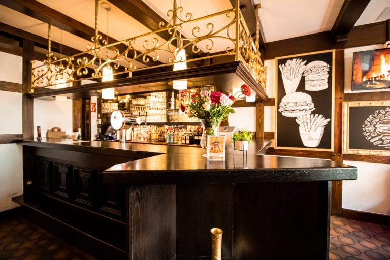 Eine gemütliche Bar mit einem dunklen Holztresen, einem dekorativen Geländer, Blumen auf dem Tresen und Regalen mit Flaschen dahinter. An den Wänden hängen gerahmte Zeichnungen von Burgern, Pommes und einem Kamin. Warme Hängelampen spenden sanftes Licht.