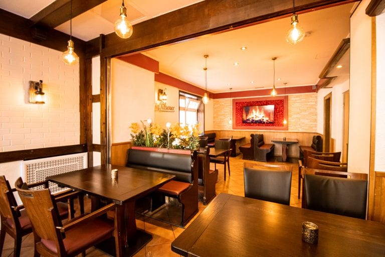 Ein gemütliches, warm beleuchtetes Restaurant mit dunklen Holztischen und -stühlen, gepolsterten Sitzbänken, dekorativen Blumen und einem digitalen Kaminbild an einer rot akzentuierten Wand. Der Raum wirkt einladend und gemütlich.