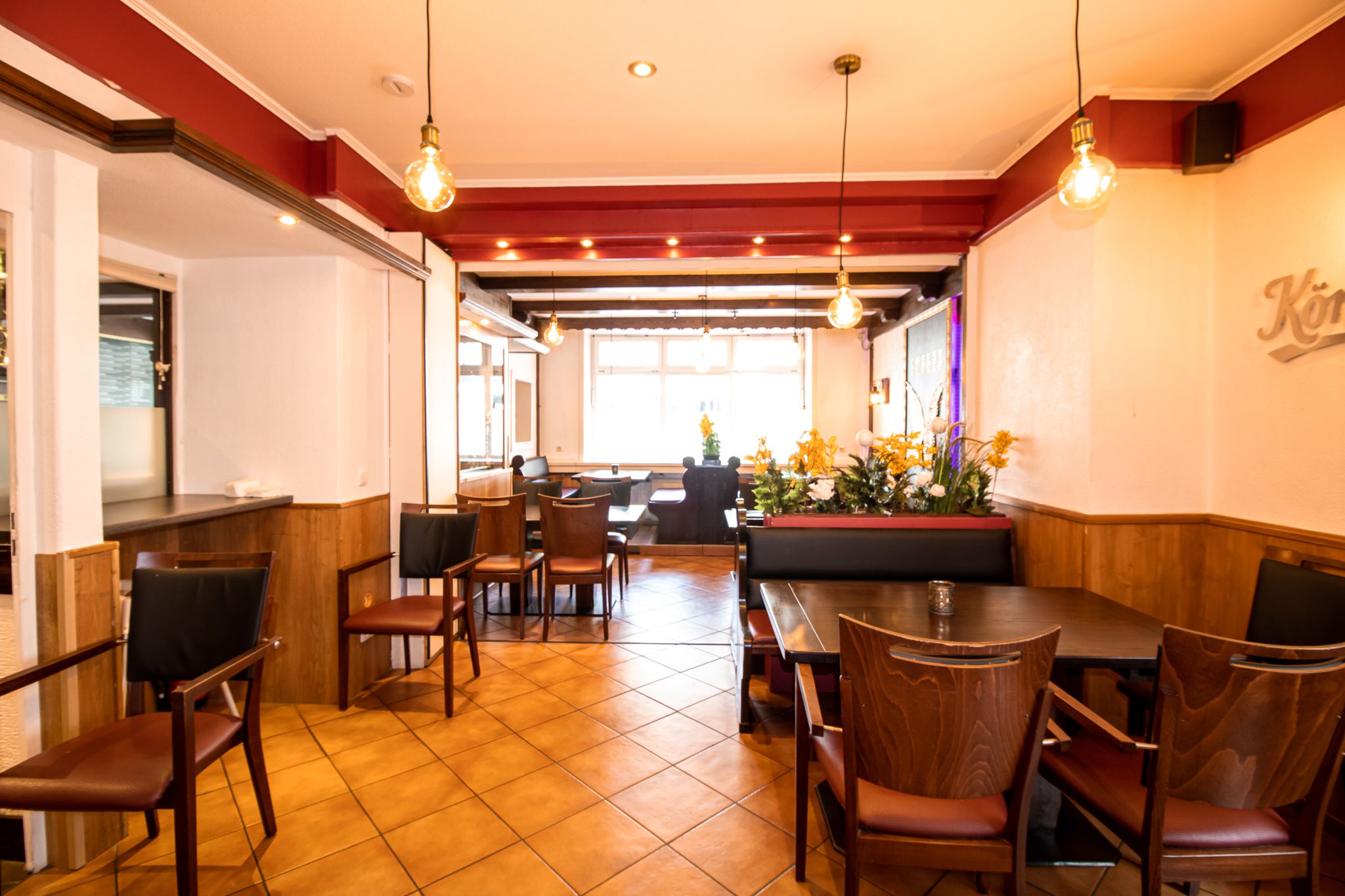 Ein helles Restaurantinterieur mit Holzstühlen und -tischen, gefliestem Boden, Hängelampen und einem Blumenarrangement, das zwei Sitzbereiche trennt. Das Sonnenlicht strömt durch große Fenster im Hintergrund.