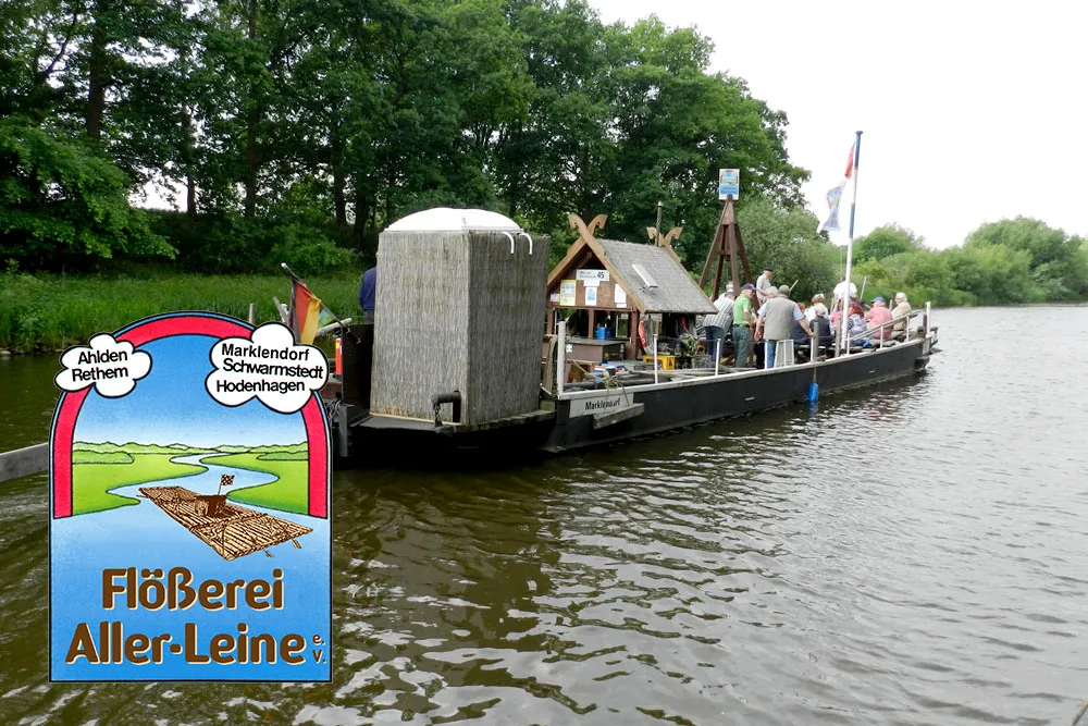 Ein Holzfloß mit mehreren Personen und einer hüttenähnlichen Konstruktion schwimmt auf einem Fluss in der Nähe grüner Bäume. Im Vordergrund steht ein illustriertes Schild mit der Aufschrift 