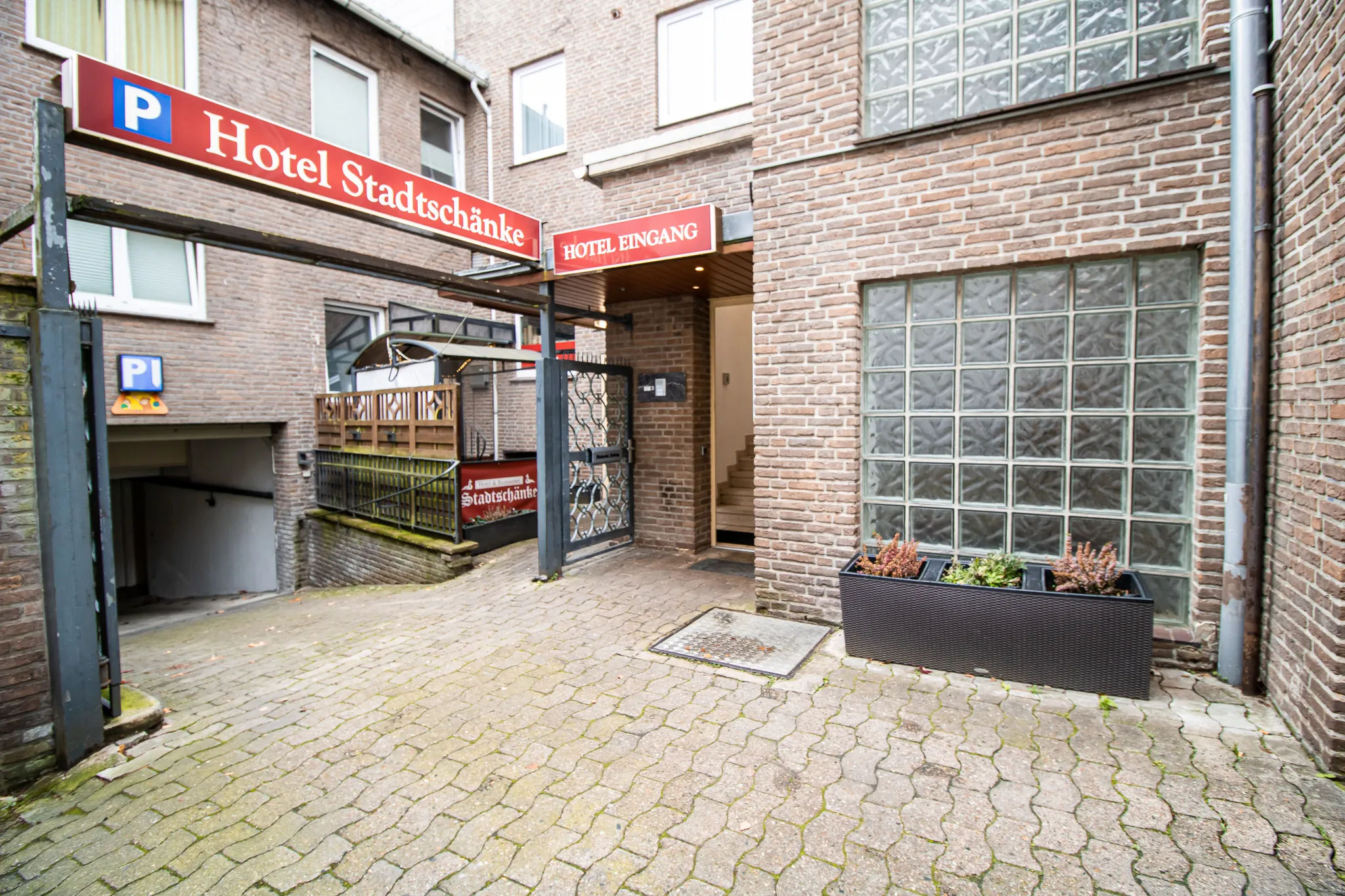 Eingang zum Hotel Stadtschänke, mit einem Schild darüber, Backsteinmauern, Glasbausteinfenstern, einer Rampe, die zur Tiefgarage führt, und einem Pflanzkübel mit kleinen Pflanzen auf einem gepflasterten Innenhof.