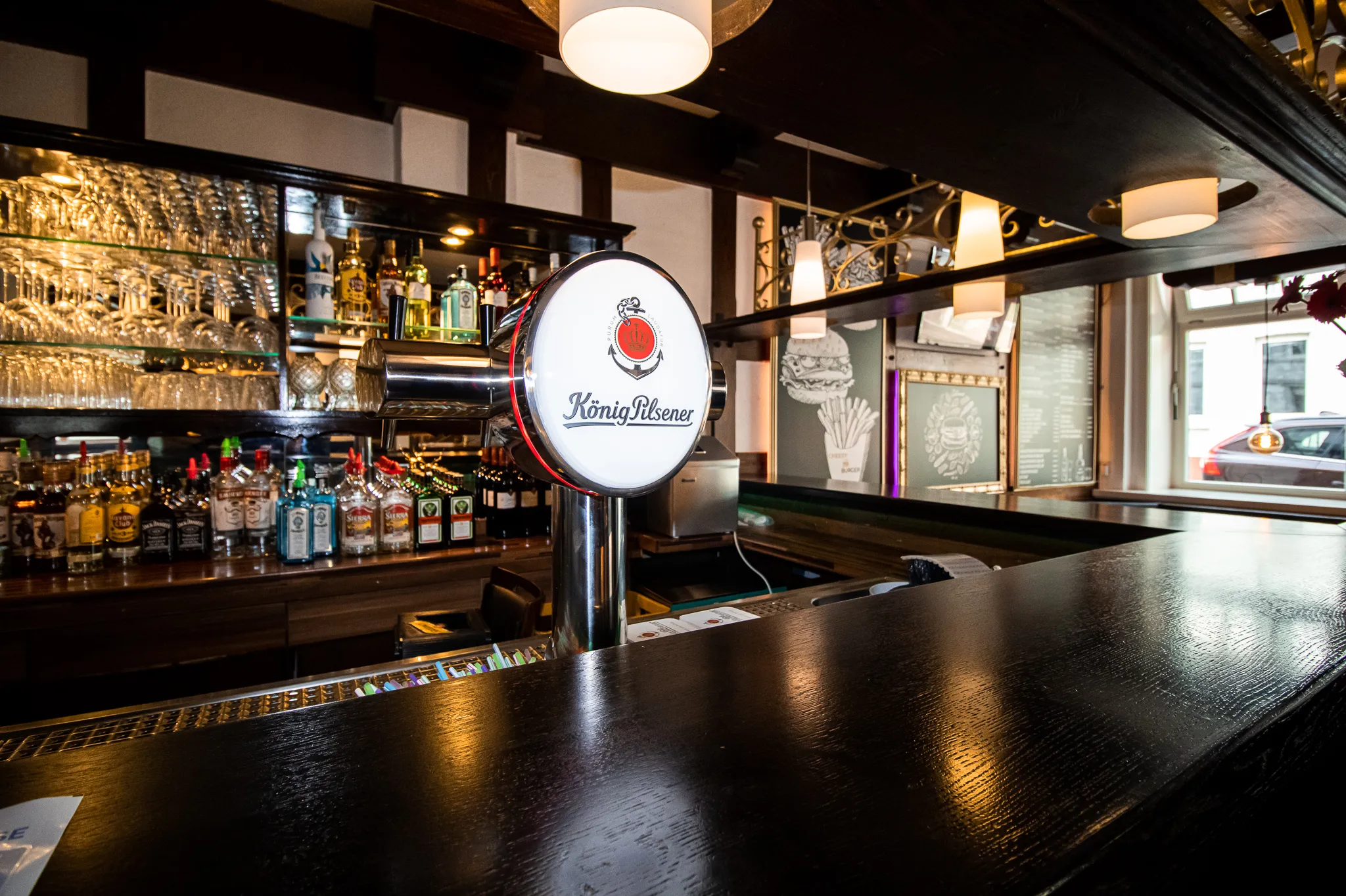 Eine gut beleuchtete Bar mit einem König-Pilsener-Zapfhahn in der Mitte, Regalen mit Spirituosenflaschen und Gläsern hinter der Bar und einem großen Fenster auf der rechten Seite, das die Straße draußen zeigt.