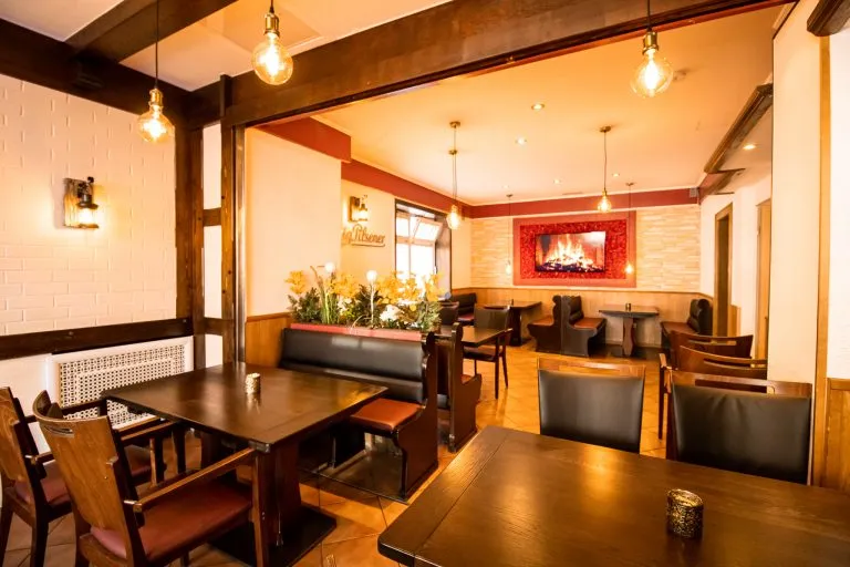 Ein gemütliches, warm beleuchtetes Restaurant mit dunklen Holztischen und -stühlen, gepolsterten Sitzbänken, dekorativen Blumen und einem digitalen Kaminbild an einer rot akzentuierten Wand. Der Raum wirkt einladend und gemütlich.