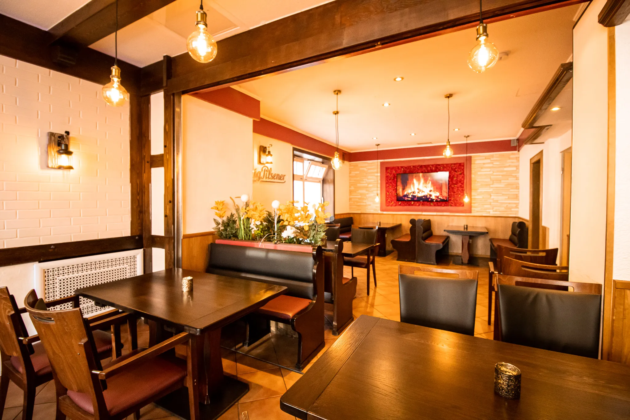 Ein gemütliches, warm beleuchtetes Restaurant mit dunklen Holztischen und -stühlen, gepolsterten Sitzbänken, dekorativen Blumen und einem digitalen Kaminbild an einer rot akzentuierten Wand. Der Raum wirkt einladend und gemütlich.