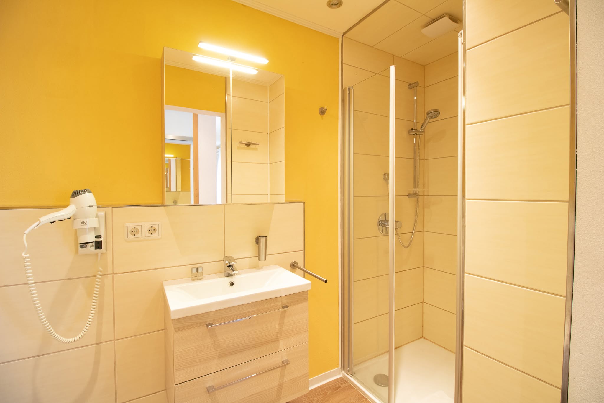 Modernes Badezimmer mit gelben Wänden, einem Spiegel über einem Waschbecken mit Unterschrank, einem eingebauten Haartrockner und einer verglasten Dusche mit Regenbrause und beigen Fliesen.