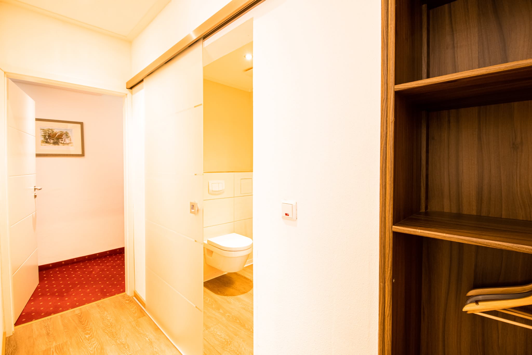 Ein heller Flur mit Holzfußboden führt zu einem Badezimmer mit Schiebetür. Das Bad ist mit einer weißen Toilette ausgestattet. Auf der rechten Seite befindet sich ein Holzregal mit ein paar Gegenständen auf der unteren Ablage.