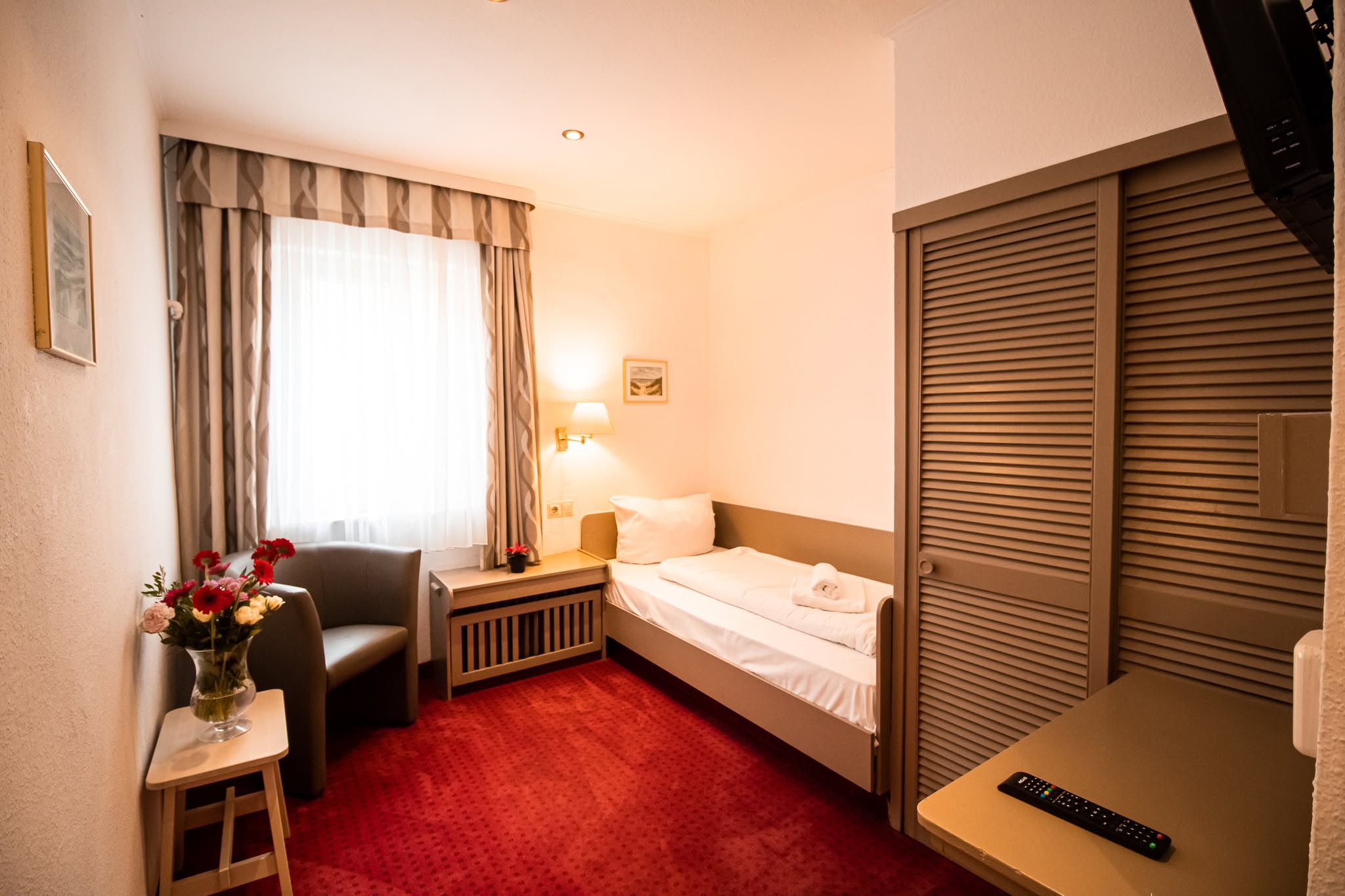 Ein gemütliches Hotelzimmer mit einem Einzelbett, rotem Teppich, einem kleinen Tisch mit Blumen, einem Sessel, einem Fenster mit gestreiften Vorhängen und einer Fernsehfernbedienung auf einer Kommode. Das Zimmer ist warm beleuchtet und ordentlich eingerichtet.