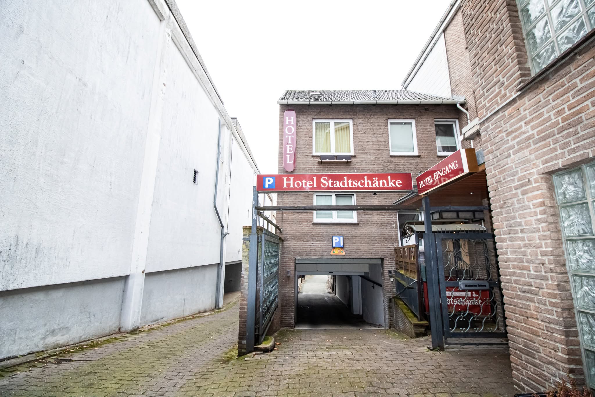 Eine schmale Gasse führt zu einer Tiefgarageneinfahrt für das Hotel Stadtschänke, mit Schildern für Parkplatz und Hoteleingang an einem Backsteingebäude. Die Umgebung besteht aus weißen und braunen Mauern und einem gepflasterten Gehweg.