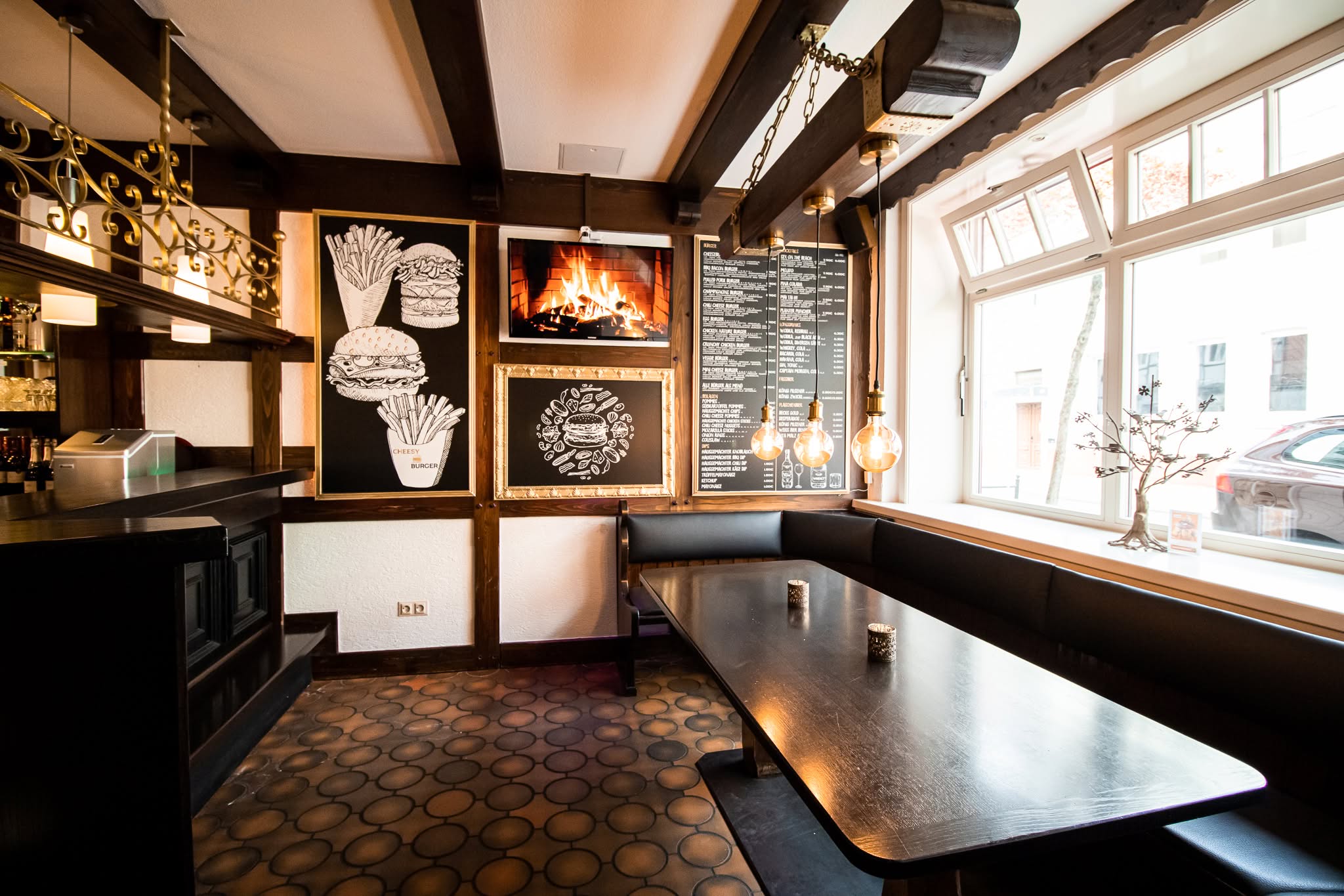 Ein gemütliches Restaurant mit einer schwarzen gepolsterten Bank, einem dunklen Holztisch, Wandbildern von Burgern und Pommes frites, einem Kaminbild, Hängelampen und einer großen Speisekarte an einem hellen Fenster.