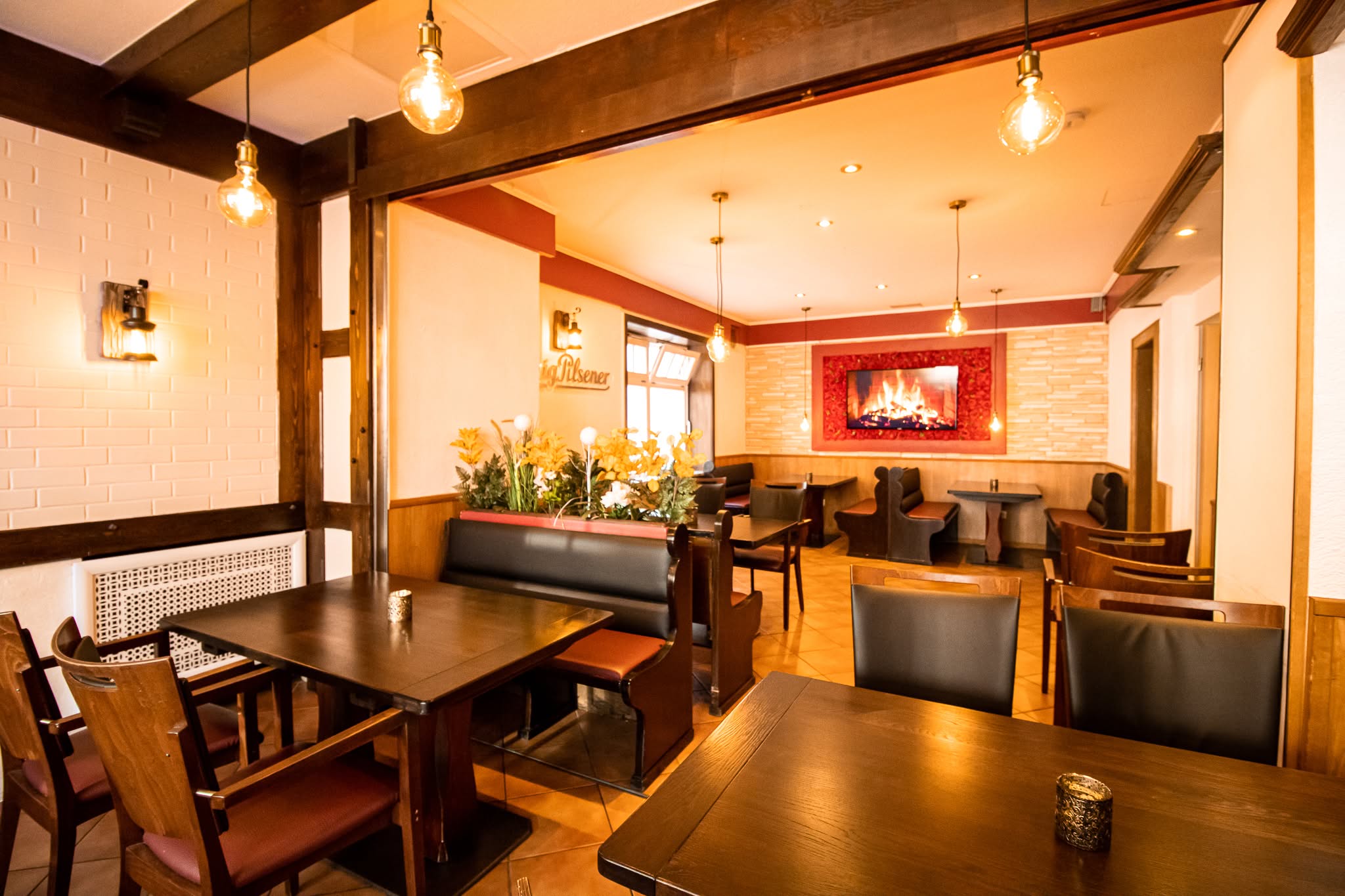 Ein gemütliches, warm beleuchtetes Restaurant mit dunklen Holztischen und -stühlen, gepolsterten Sitzbänken, dekorativen Blumen und einem digitalen Kaminbild an einer rot akzentuierten Wand. Der Raum wirkt einladend und gemütlich.